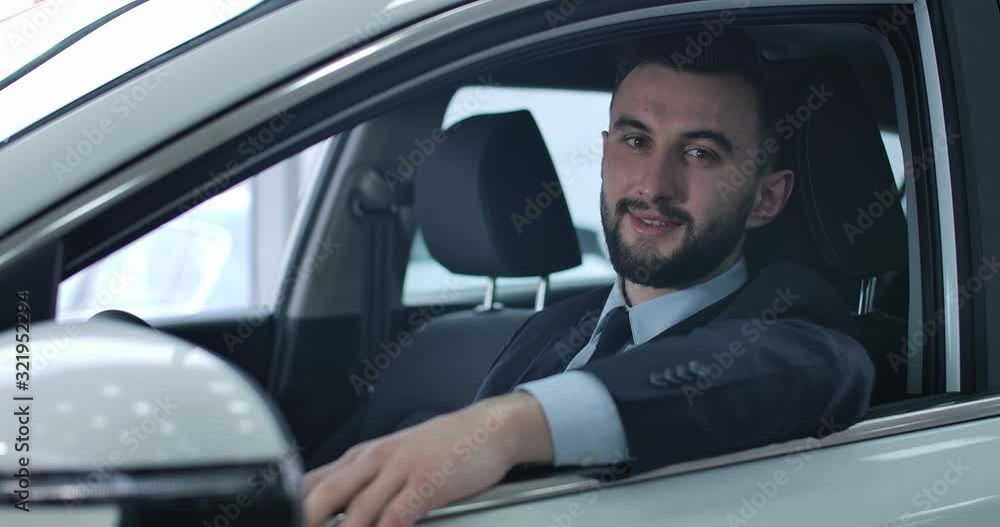 Close-up portrait of satisfied Caucasian young man posing in car on driver's seat. Positive handsome man buying new automobile in dealership. Cinema 4k ProRes HQ. Close-up portrait of satisfied Caucasian young man posing in car on driver's seat. Positive handsome man buying new automobile in dealership. Cinema 4k ProRes HQ.