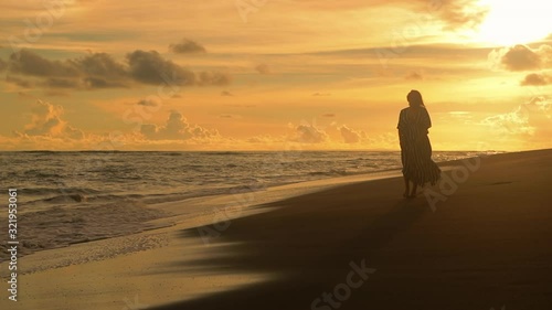 Young sexy woman in black dress walking and looking at the sunset at the sand beach. Pretty girl walking towards ocean. Femininity concept. City people at vacation. Business woman at summer vacation.