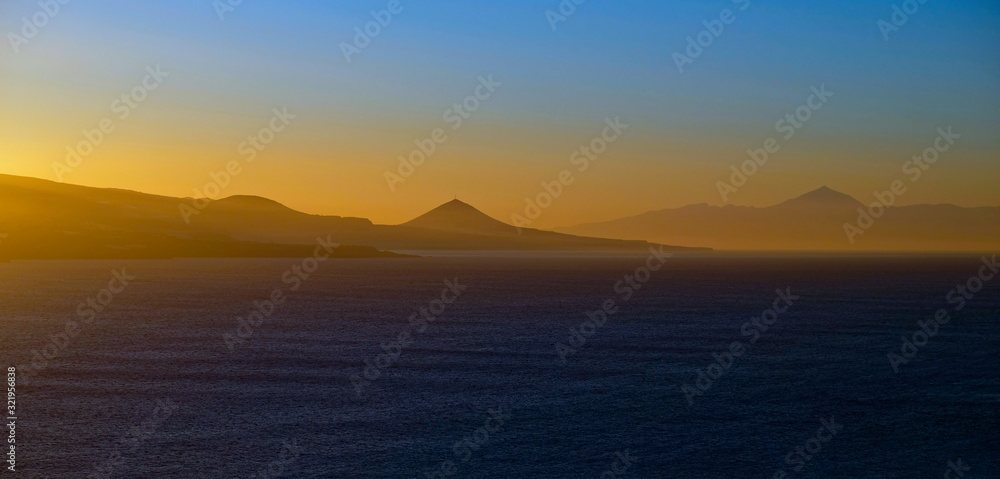 Spectacular sky colors and sunset landscape of a volcanic island with sun reflections in calm ocean.