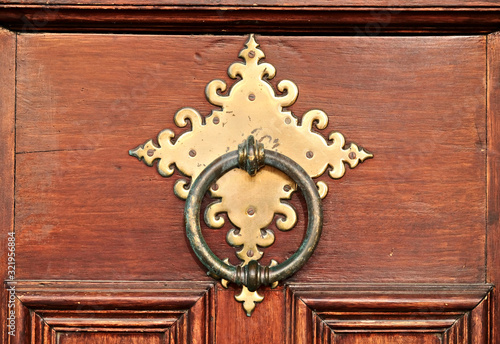 Large round heavy metal door knocker with an ornamental metal plate on warm brown varnished wooden door.