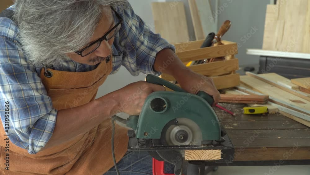 4K Professional Asian senior craftsman carpenter using carpentry chainsaw cutting plank of wood for production of modern wooden furniture in studio workshop. Retirement lifestyle DIY working concept. 4K Professional Asian senior craftsman carpenter using carpentry chainsaw cutting plank of wood for production of modern wooden furniture in studio workshop. Retirement lifestyle DIY working concept.