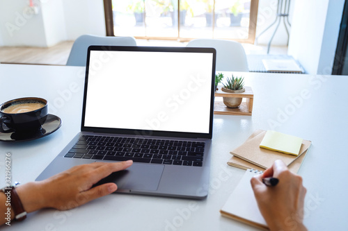 Mockup image of a woman using and touching laptop touchpad with blank white desktop screen while writing on a notebook