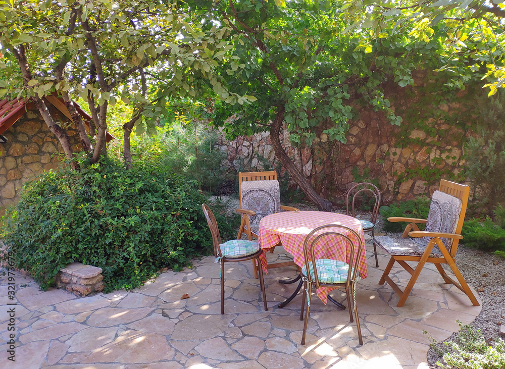 Summer, relaxation area in retro style. Round table, Vienna chairs, 2 chairs with wooden armrests. Good sunny day, outdoors. Natural stone, a shadow from the foliage of trees, a cozy beautiful place.