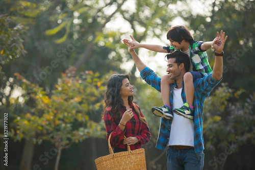 Happy young couple standing in a garden with the man carrying their son on his shoulders and the woman carrying a basket.