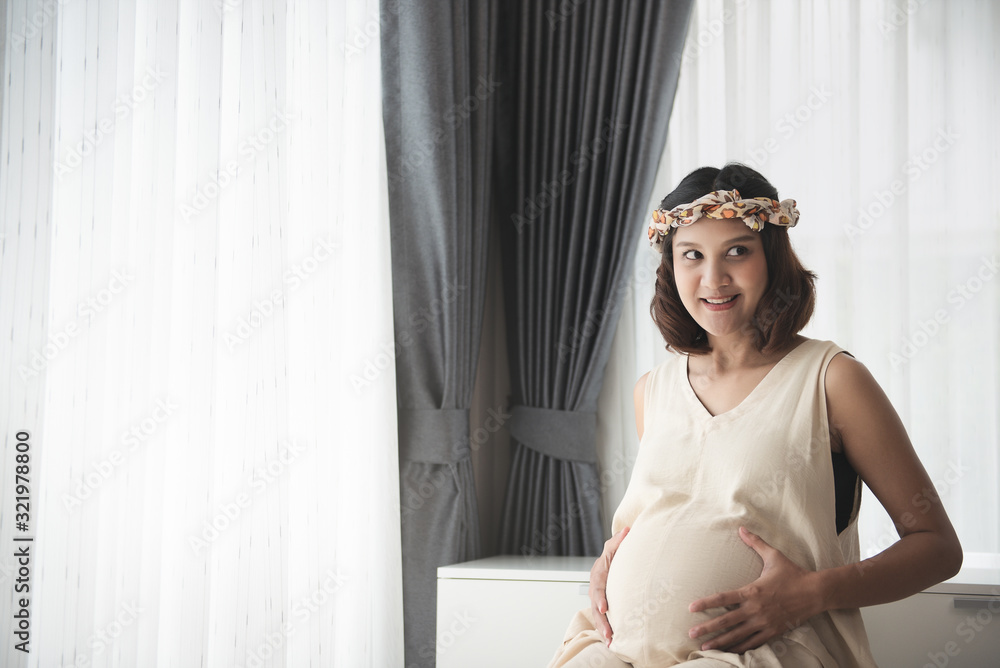 Pregnancy, Pregnant woman touch big belly in Maternity Dresses sitting near window at home. happiness of motherhood preparation and expectation with copy space concept.