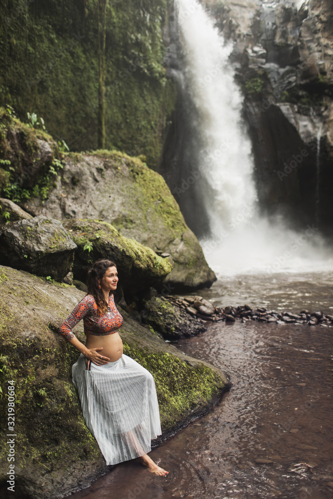 Young authentic Spanish pregnant woman enjoying amazing cascade waterfall in Ubud. Harmony with nature, waiting baby.