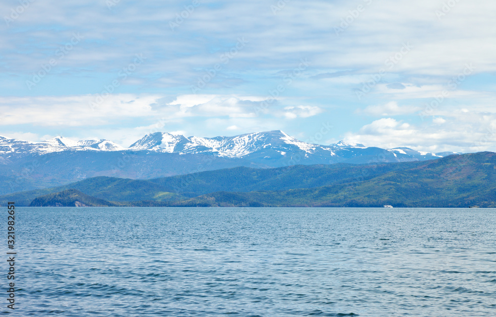 Beautiful natural background with snowy mountain peaks and forested hills on the shore of the Chivyrkuisky Bay of Lake Baikal on a June day