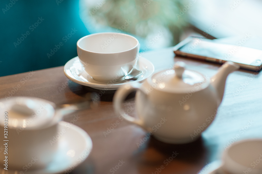 White cups and a white teapot on a table in a cafe. A mobile phone lies next to the cup. White cups and a white teapot on a table in a cafe. A mobile phone lies next to the cup.