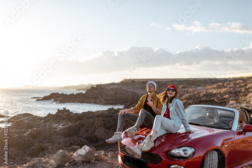 Young lovely couple enjoying landscapes, sitting together on a car hood, traveling by car on the rocky ocean coast. Carefree lifestyle, love and travel concept