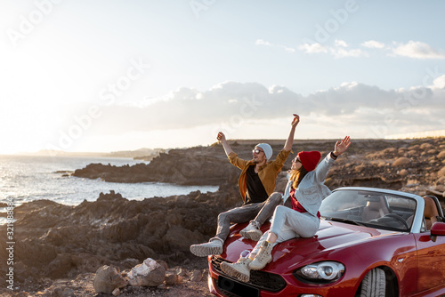 Young lovely couple enjoying landscapes, sitting together on a car hood, traveling by car on the rocky ocean coast. Carefree lifestyle, love and travel concept