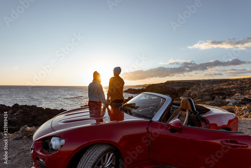 Couple enjoying beautiful views on the ocean, hugging together near the car on the rocky coast, wide view from the side with copy space on the sky