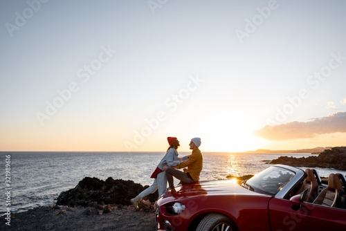 Couple enjoying beautiful views on the ocean, hugging together near the car on the rocky coast, wide view from the side with copy space on the sky