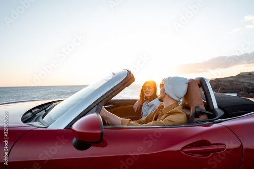 Konstfotografi Joyful couple enjoying vacations, driving together convertible car near the ocean on a sunset