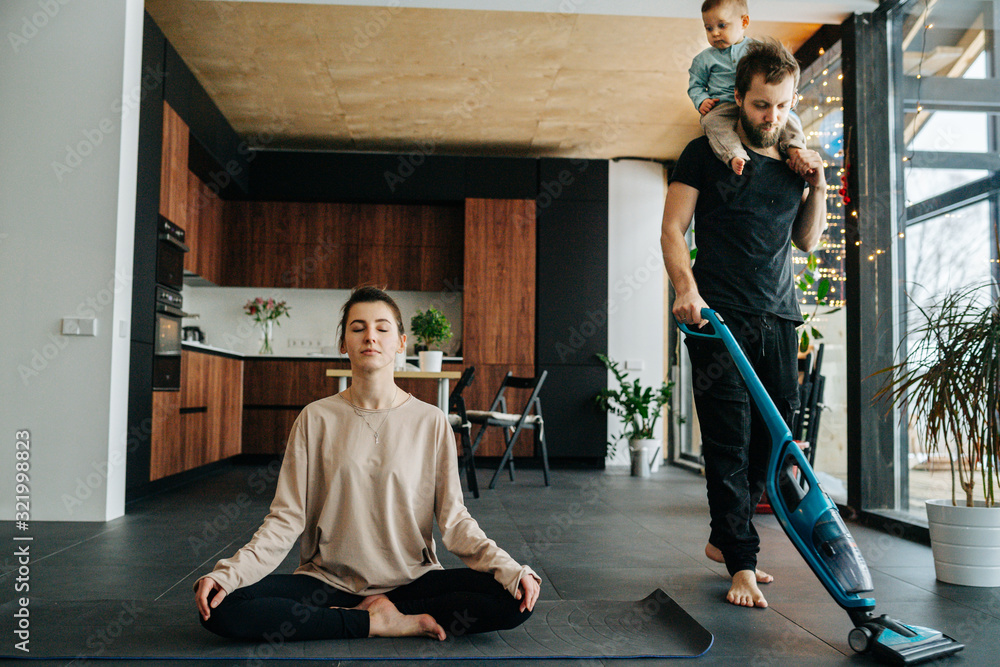 Obraz premium Mother sits at easy pose, while father vacuums floor with baby riding on neck