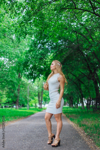 Portrait of a charming blond woman wearing beautiful white dress standing on the road under trees.