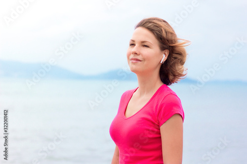 Happy pensive girl, young beautiful woman listening to music in wireless new modern earphones, enjoying song, good weather, breath deep fresh air, dreaming outdoor. Sea background, lady in red T-shirt © Евгений Шемякин