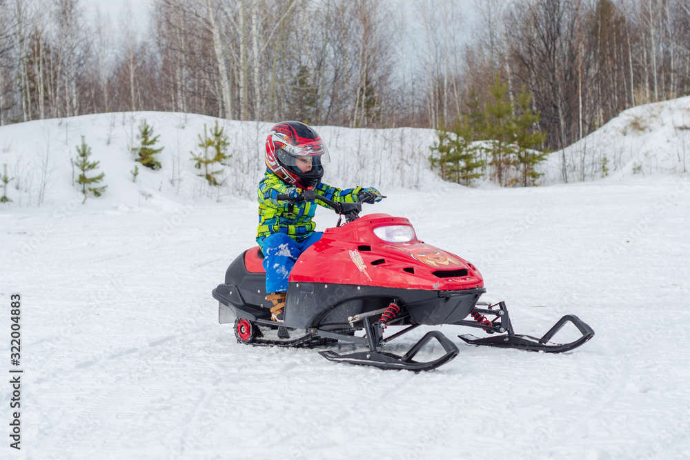 a child in a protective helmet rides a red snowmobile in winter