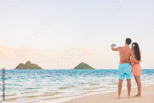 Beach vacation tourists couple taking picture with phone of Hawaii islands. Honeymoon summer travel holidays.