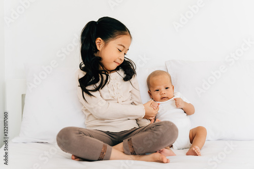 Caring Asian girl in casual wear with crossed legs holding hand of small brother sitting on bed in house