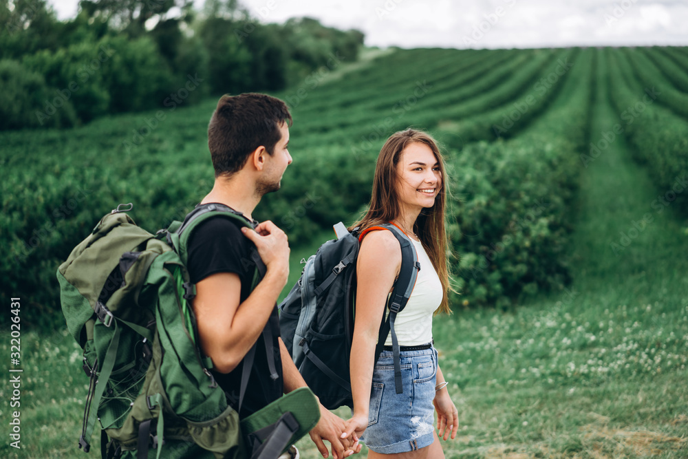 Portrait of happy young couple with backpacks on the field in spring. Man and woman walking in currant plantations