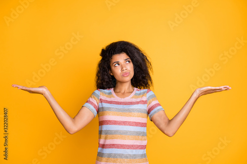 Portrait of her she nice attractive doubtful wavy-haired girl in striped tshirt holding on palms invisible objects weighing thinking isolated on bright vivid shine vibrant yellow color background photo