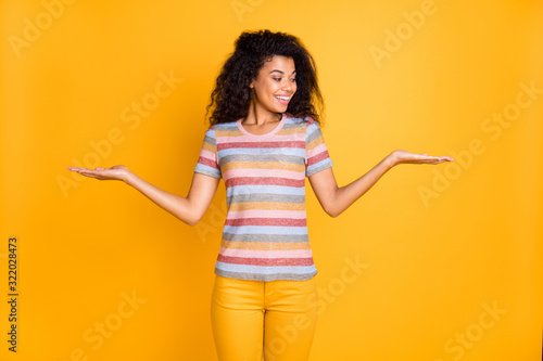 Portrait of her she nice attractive cheerful cheery wavy-haired girl in striped tshirt holding on palms two invisible objects weighing isolated on bright vivid shine vibrant yellow color background