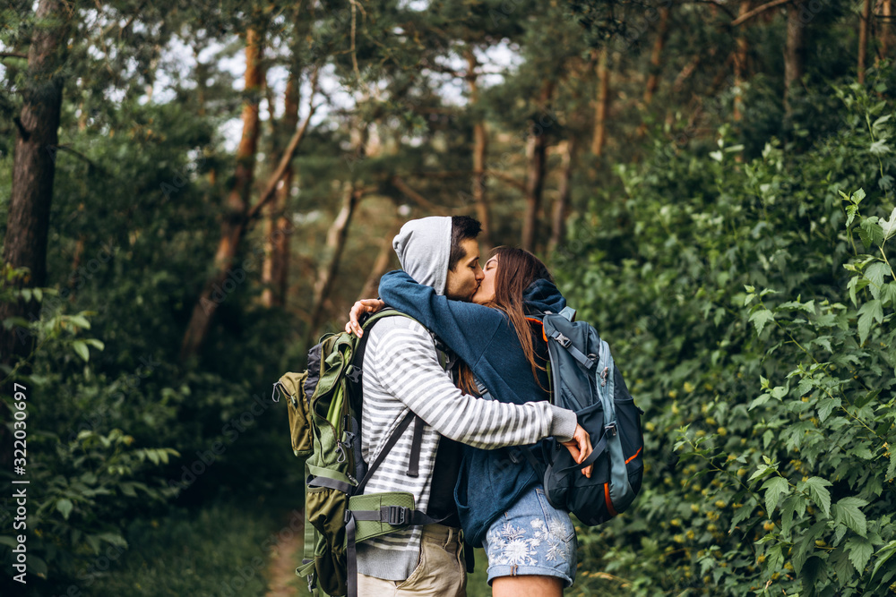 Young couple with backpacks on their backs in the forest. Loving man kisses his beautiful girlfriend on a hike in the woods