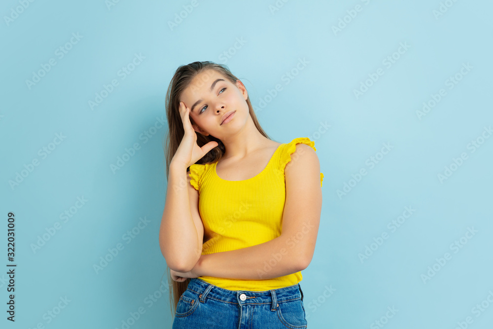 Thoughtful. Caucasian teen girl's portrait isolated on blue background. Beautiful model in casual yellow wear. Concept of human emotions, facial expression, sales, ad. Copyspace. Looks cute. Thoughtful. Caucasian teen girl's portrait isolated on blue background. Beautiful model in casual yellow wear. Concept of human emotions, facial expression, sales, ad. Copyspace. Looks cute.