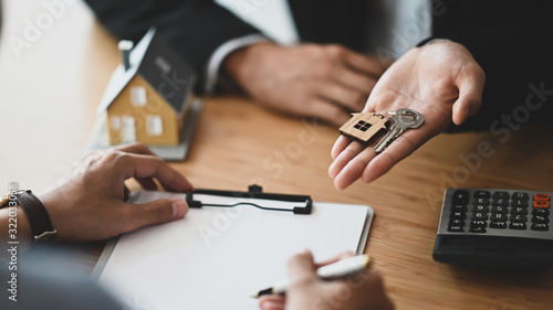 Cropped shot of Real estate agent giving a property key to his customer while signing an agreement on clipboard at the modern wooden table. Realtor/Broker/Seller/Dealer concept. photo