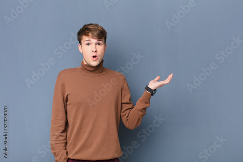 young blonde man looking surprised and shocked, with jaw dropped holding an object with an open hand on the side isolated against flat wall photo