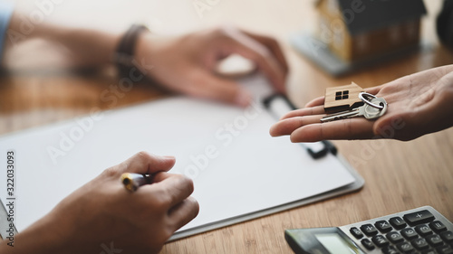 Cropped shot of Real estate agent giving a property key to his customer while signing an agreement on clipboard at the modern wooden table. Realtor/Broker/Seller/Dealer concept.