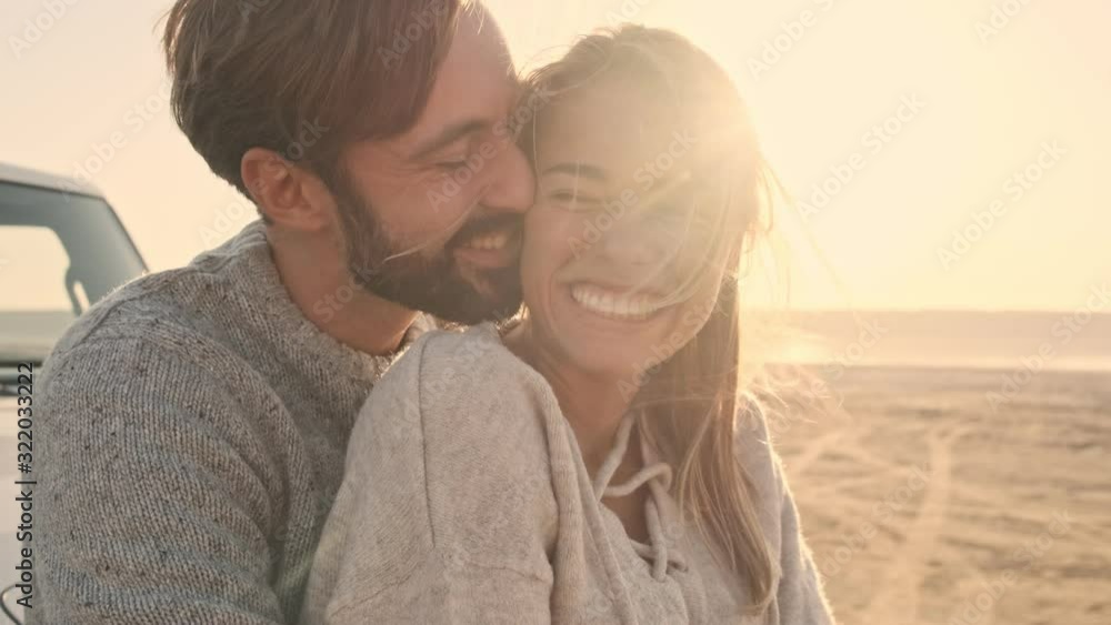 A young couple in love man and woman are hugging and kissing while standing near the suv car on the lake in summer A young couple in love man and woman are hugging and kissing while standing near the suv car on the lake in summer
