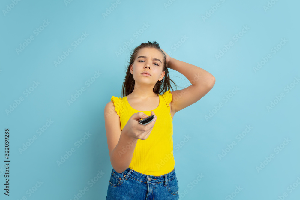 Using TV remote. Caucasian teen girl's portrait isolated on blue background. Beautiful long hair model in casual. Concept of human emotions, facial expression, sales, ad. Copyspace. Looks cute. Using TV remote. Caucasian teen girl's portrait isolated on blue background. Beautiful long hair model in casual. Concept of human emotions, facial expression, sales, ad. Copyspace. Looks cute.