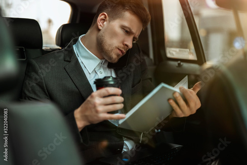 successful businessman drinks coffee, getting ready for important negotiations while riding on the back seat of car on way to meetting with partners,, multitasking concept.