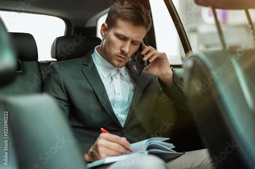 successful businessman takes notes to planner while having business phone conversation riding on back seat of car on way to meetting with partners, multitasking concept © studioprodakshn