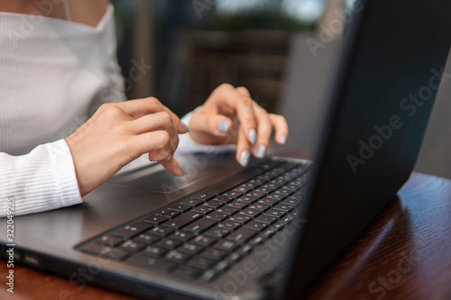 Beautiful young woman in white t-shirt is working on laptop and smiling while sitting outdoors in cafe. Young female using laptop for work. Female freelancer working on laptop in an outdoor cafe.