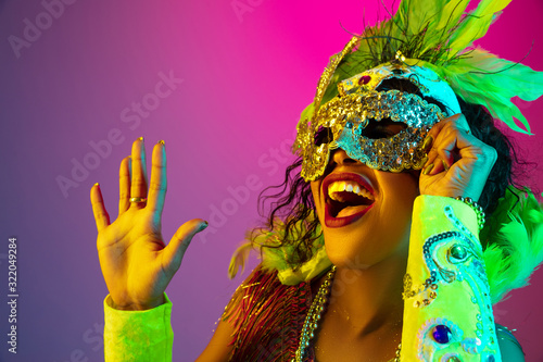 Close up. Beautiful young woman in carnival, stylish masquerade costume with feathers on gradient background in neon light. Concept of holidays celebration, festive time, dance, party, having fun. photo