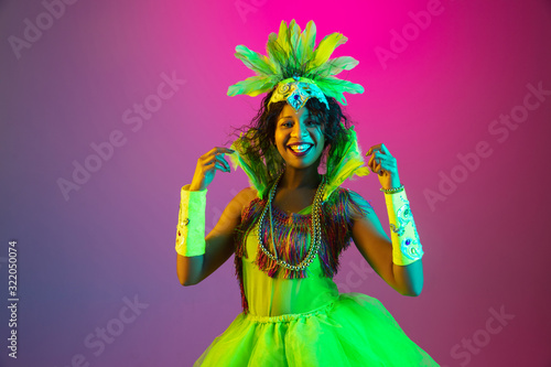 Bright. Beautiful young woman in carnival, stylish masquerade costume with feathers dancing on gradient background in neon. Concept of holidays celebration, festive time, dance, party, having fun.