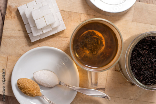 Cukier w kostkach, cukier trzcinowy, cukier biały sypki oraz miód gryczany na łyżeczce leży obok szklanki z zaparzonym naparem z ziół. 
