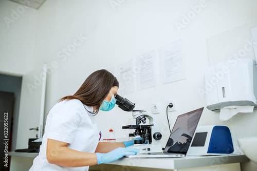 Charming caucasian laboratory assistant in white uniform, with protective mask and rubber gloves sitting in lab looking blood sample trough microscope and using laptop.