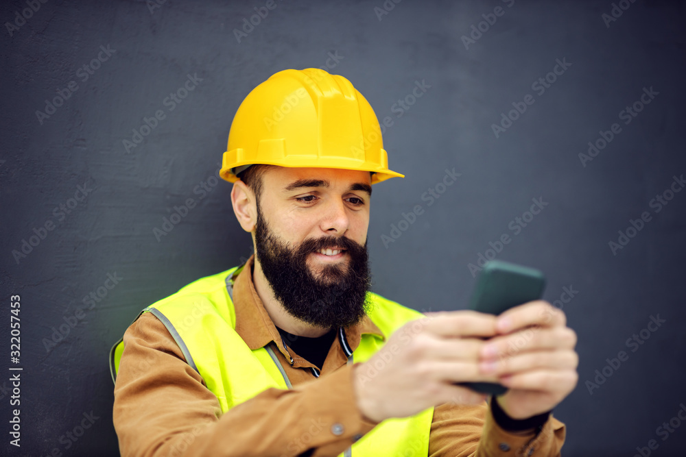 Close up of handsome smiling bearded worker in vest, with helmet on head sitting, using smart phone and taking a break from work. Close up of handsome smiling bearded worker in vest, with helmet on head sitting, using smart phone and taking a break from work.
