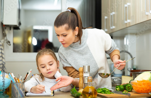 Girl writing school lesson during mother cooking at kitchen