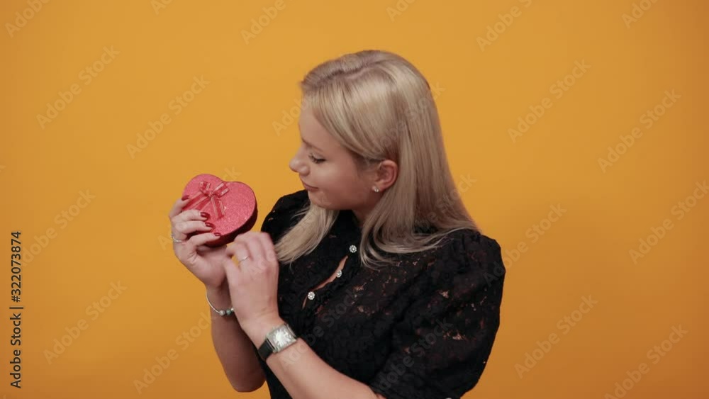 Beautiful blonde girl in black dress on yellow background a woman in love holds gift in the form of heart box Beautiful blonde girl in black dress on yellow background a woman in love holds gift in the form of heart box