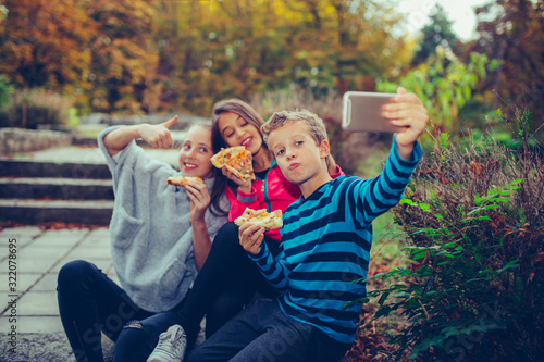 Group of a three happy children taking selfie by smart phone while eating pizza outdoors