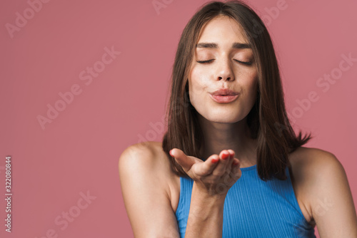 Image of young brunette woman wearing top blowing air kiss at camera