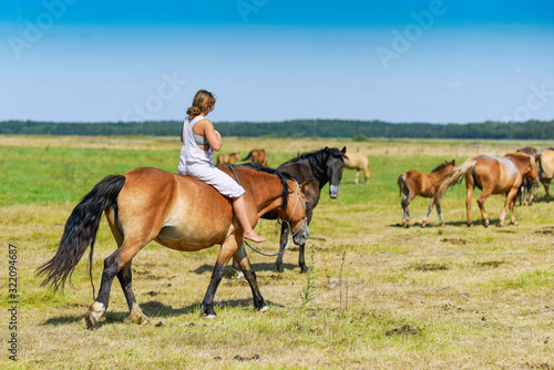 A girl rides a horse across a farm field. Photographed from behind.
