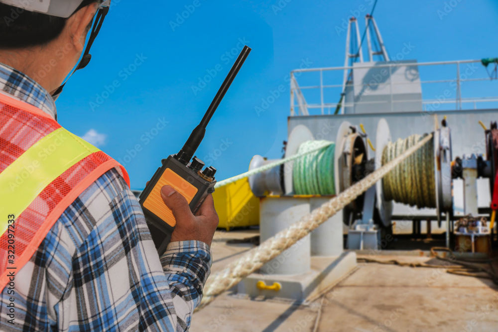 crew worker radio walkie talkie in hand on vessel, cargo ship on mooring winch, windlass with rope anchor in drum background.