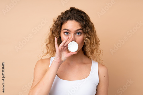Young blonde woman with curly hair isolated on beige background with moisturizer