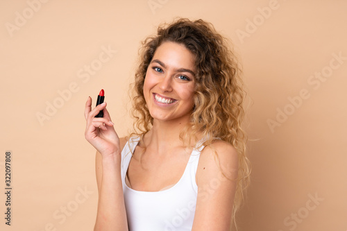 Young blonde woman with curly hair isolated on beige background holding red lipstick