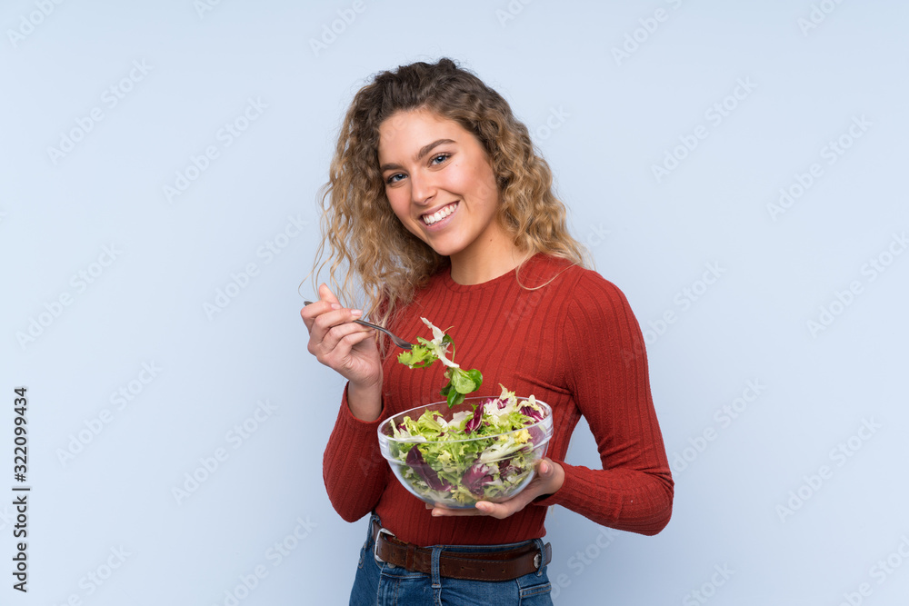 Young blonde woman with curly hair holding a salad over isolated wall
